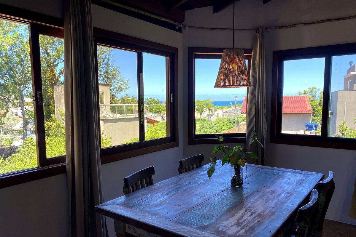 Un espace repas avec une table et des chaises en bois, de grandes fenêtres offrant une vue sur la verdure et les bâtiments, et un luminaire suspendu.