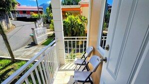 A balcony with a white door, a white chair, and a view of a street with houses and trees. - Lovely 2-Bedroom Apartment, Shared Outdoor Pool, 10 minutes walk to downtown  (Roseau)