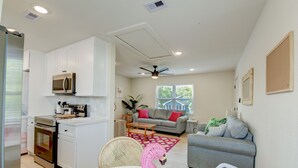 A modern kitchen with white cabinets, a microwave, and a built-in oven. There is a dining area with a white table and chairs, and a living room with a gray sofa and a red rug.