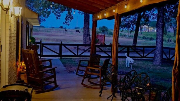 Eine Terrasse im Freien mit Holzsesseln und -tischen, ein Hund beim Spaziergang und Blick auf eine eingezäunte Weide mit Bäumen.
