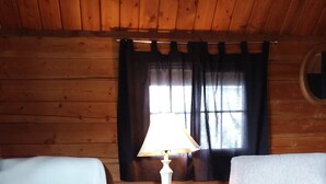 A cozy room with wooden walls and a window with curtains. There is a white lamp on a small table, a brown armchair with a decorative pillow, and a light grey armchair with a floral pillow. - Comfy cabin for two close to Grand Canyon and many Northern Arizona attractions (Williams)