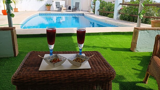 A wicker table with two glasses of red drink and bowls of food, set on a lawn with a pool and buildings in the background. - Bella Vista Apartment (Coin)