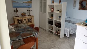A room with a dining table, chairs, a white cabinet, a bookshelf, and a bed with patterned bedding. - Bella Vista Apartment (Coin)