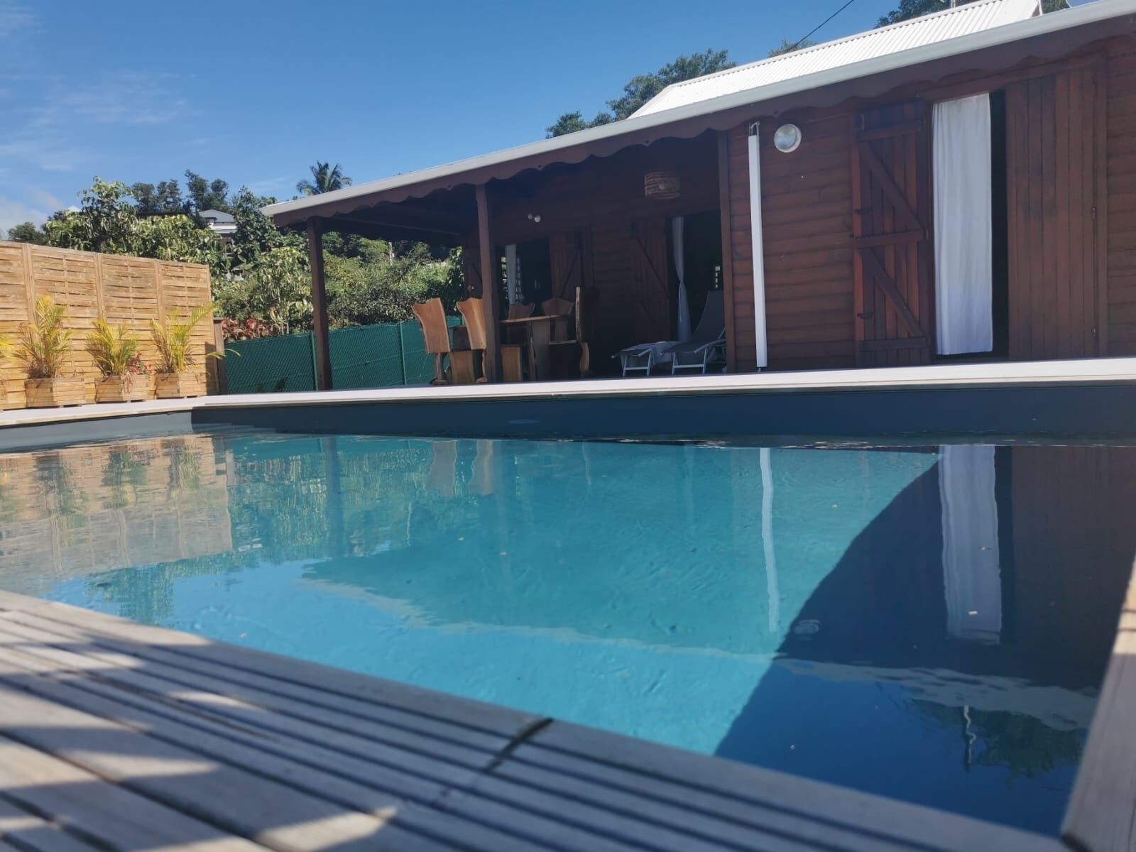 Une piscine avec une terrasse en bois, un patio couvert et une vue sur un immeuble d’habitation en arrière-plan.