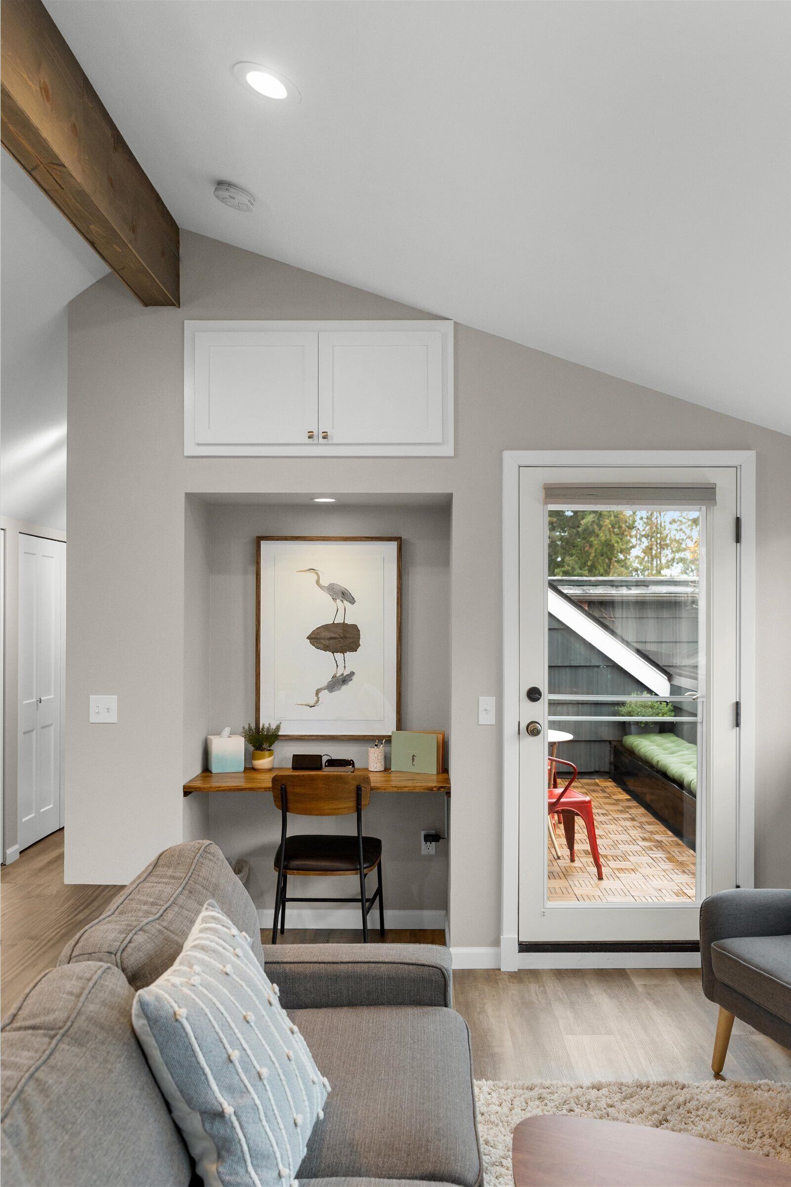 A modern living room with a gray sofa, a wooden coffee table, a framed artwork above a desk, and a sliding glass door leading to a balcony.