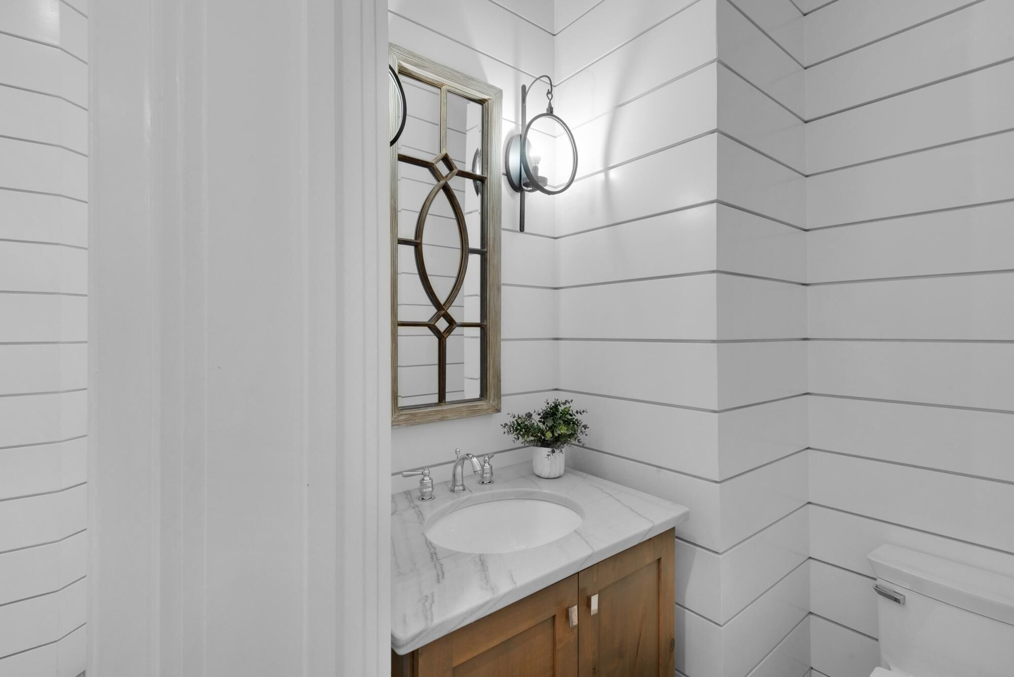 A bathroom with a white vanity, a marble sink, a wooden cabinet, a mirror with a decorative frame, and a wall-mounted light fixture.