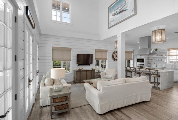 A spacious living room with a large sofa, a coffee table, and a TV. There is a kitchen area with bar stools and a dining table.