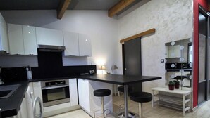 A modern kitchen with white cabinets, a black countertop, and stainless steel appliances. There are three black bar stools and a small table with plants. - Le Rivat - COMMUNAY - Gîte (COMMUNAY)