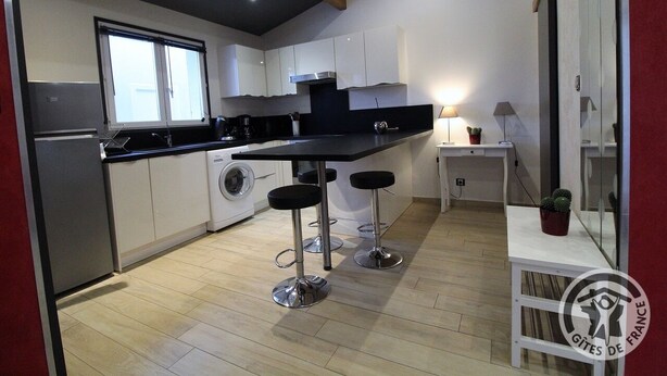 A modern kitchen with white cabinets, a black countertop, and stainless steel appliances. There are two bar stools and a small table with a lamp. - Le Rivat - COMMUNAY - Gîte (COMMUNAY)