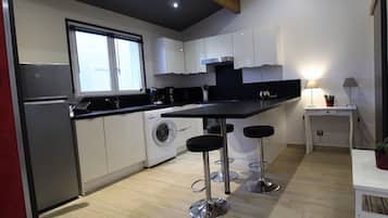 Una cocina moderna con armarios blancos, encimera negra y electrodomésticos de acero inoxidable. Hay dos taburetes de bar y una pequeña mesa con una lámpara.