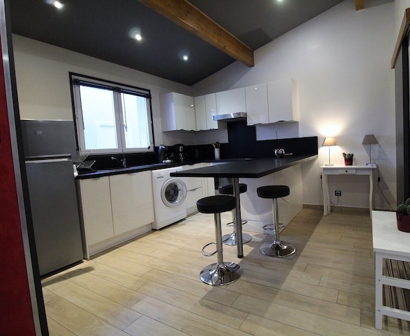 A modern kitchen with white cabinets, a black countertop, and stainless steel appliances. There are two bar stools and a small table with a lamp.