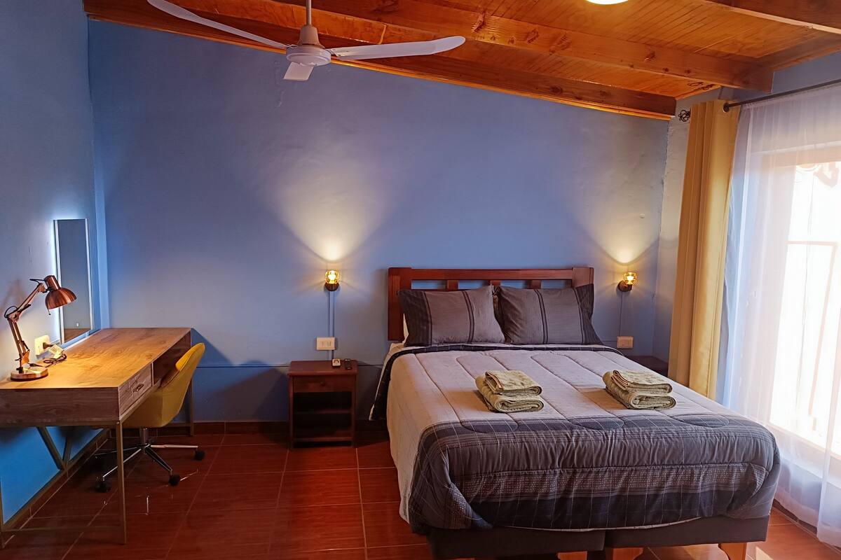 A bedroom with a wooden ceiling, a bed with a dark quilt, two bedside lamps, a desk with a lamp, and a chair.