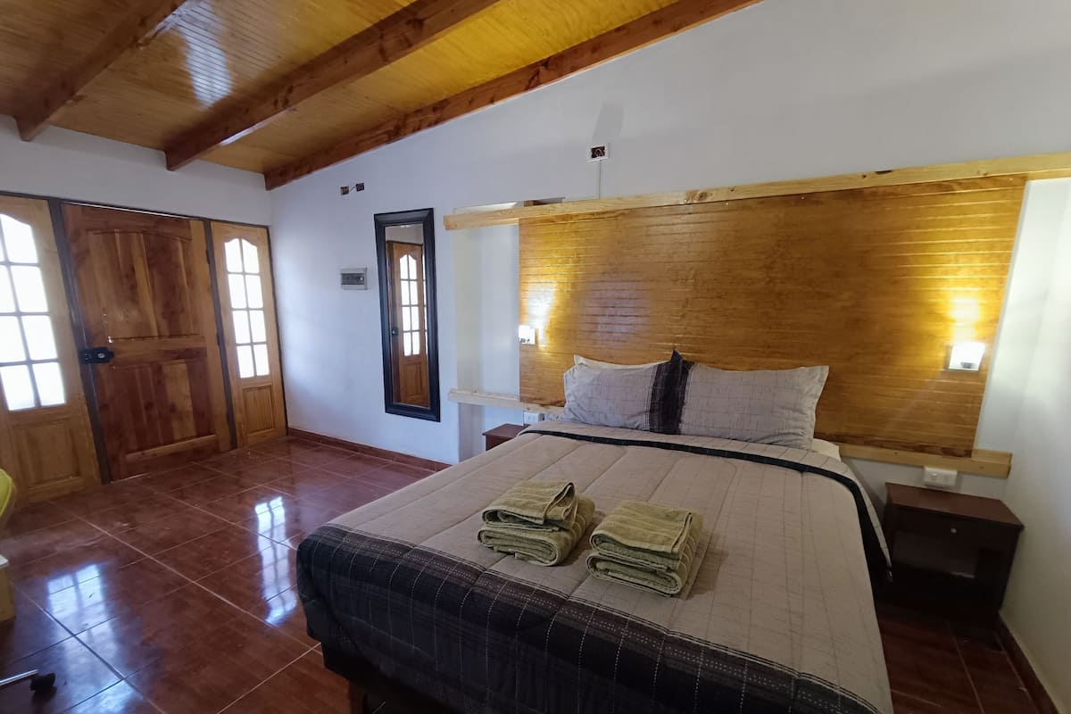 A bedroom with a wooden headboard, a bed with a quilt and towels, a nightstand, and a wooden door.