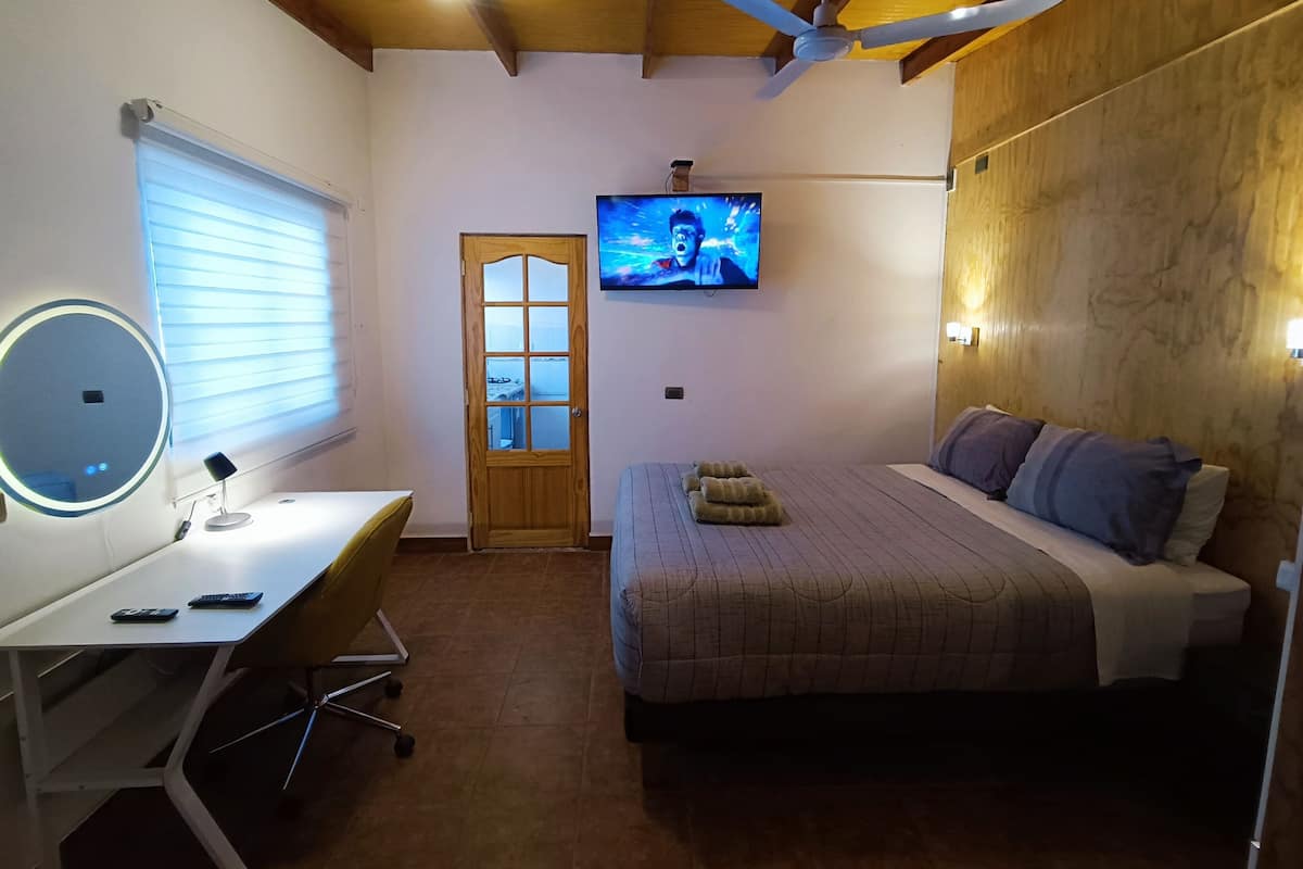 A bedroom with a bed, a desk with a chair, a television mounted on the wall, and a ceiling fan.