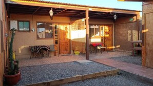 A covered outdoor seating area with wooden tables and chairs, a wooden bench, and a cactus plant. - Yurak Apart Hotel (San Pedro de Atacama)
