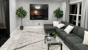 Ein modernes Wohnzimmer mit einem großen Flachbildfernseher, einem dunkelgrauen Sofa, zwei Topfpflanzen und einem Glastisch.