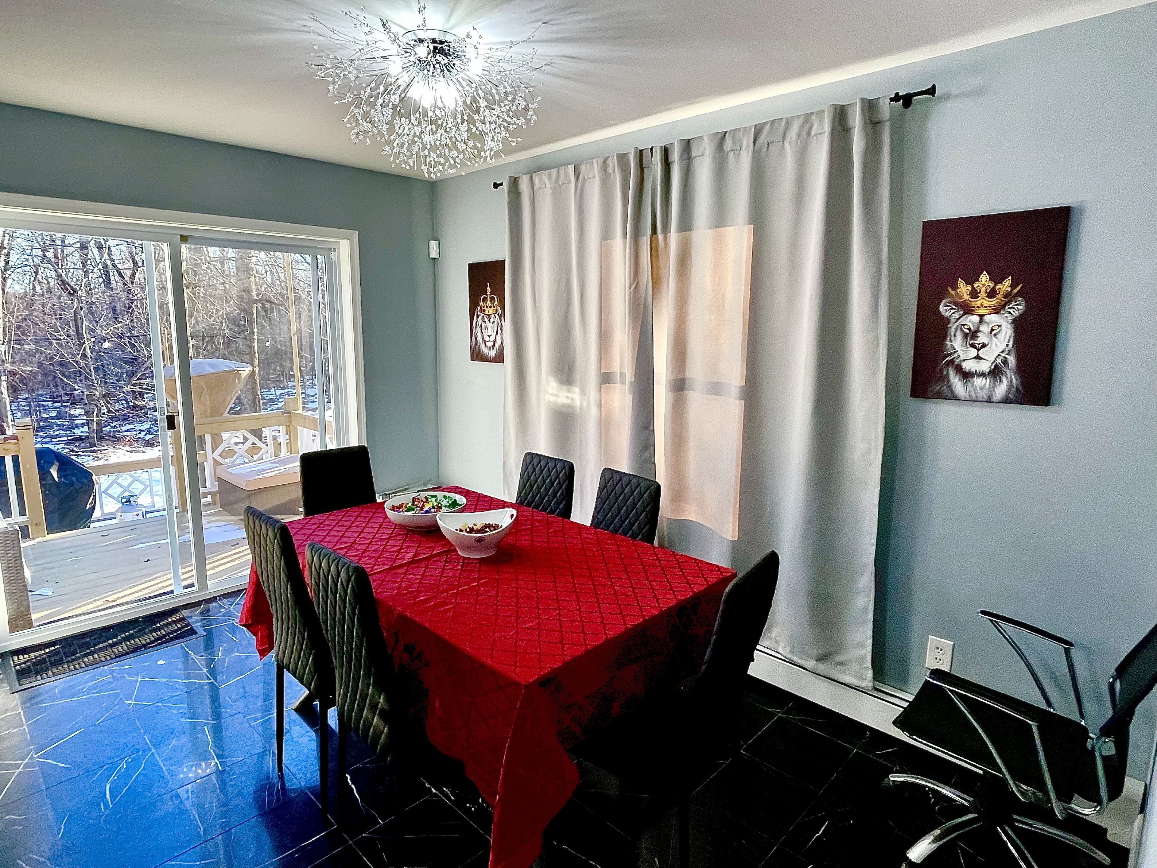 A dining area with a red tablecloth, black chairs, a chandelier, and a view of a snowy landscape through a glass door.