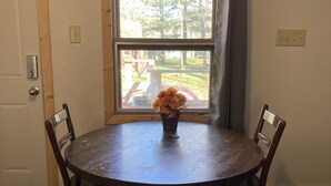 A round wooden table with four chairs, a vase with flowers, and a window with a view of a deck and trees. - Musky Joes Twin Pines Resort (Hayward)