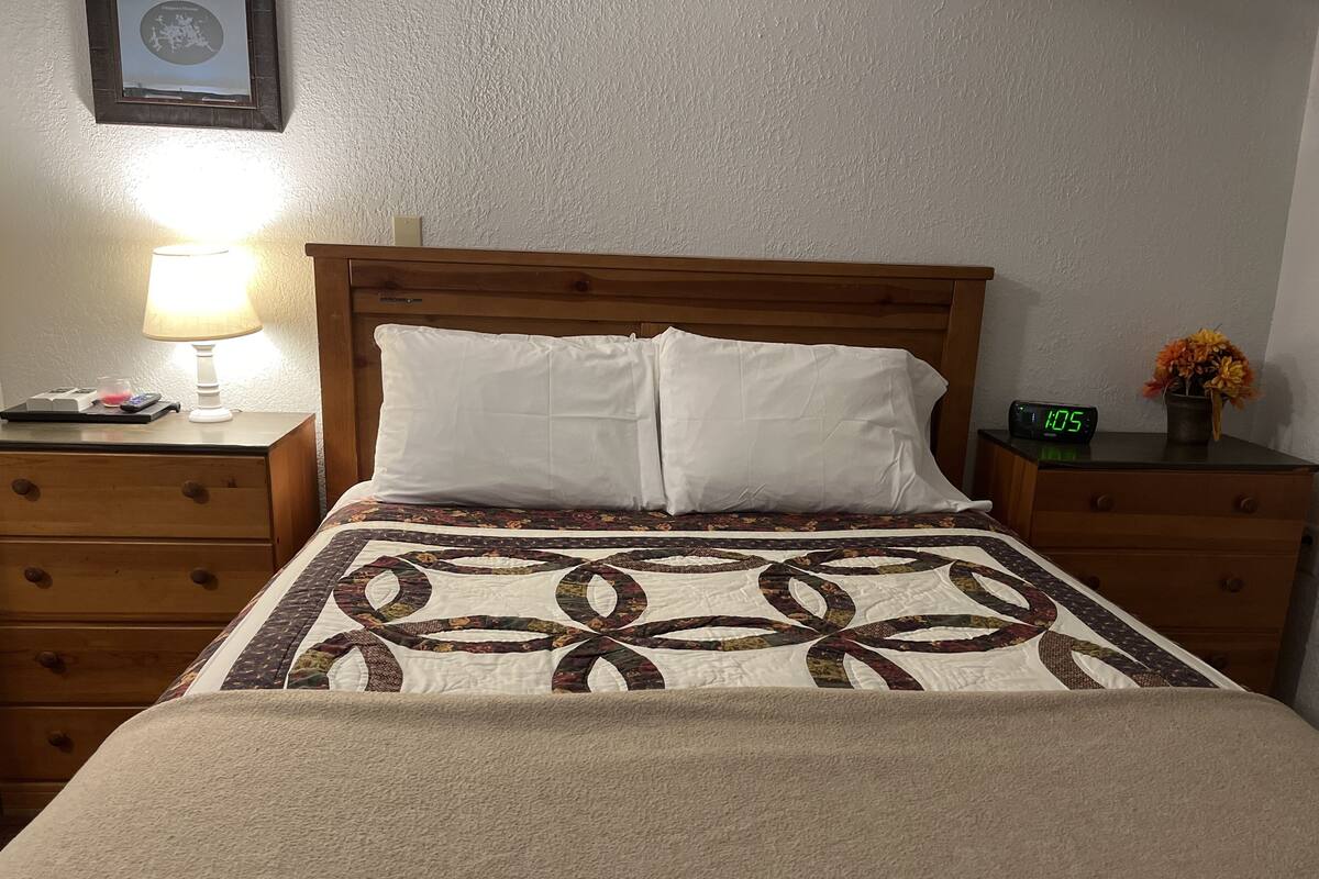 Ein ordentlich bezogenes Bett mit einer gemusterten Steude, einem hölzernen Kopfteil und einem Nachttisch mit Lampe und Uhr.