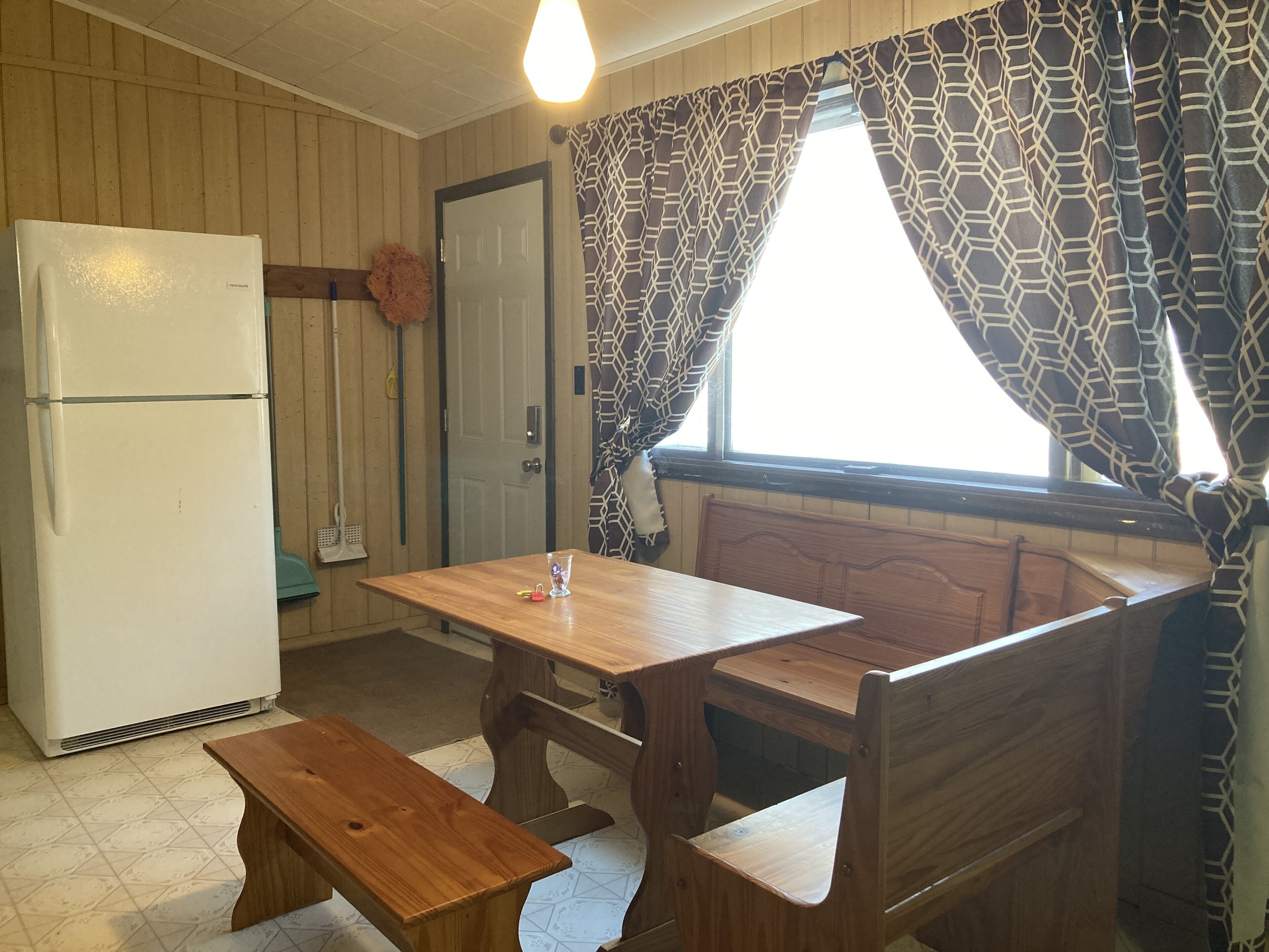 Une cuisine de taille réduite, avec une table et un banc en bois, un réfrigérateur blanc et des rideaux à motifs.