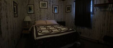 A dimly lit room with a bed, a nightstand, a lamp, and framed pictures on the wall.