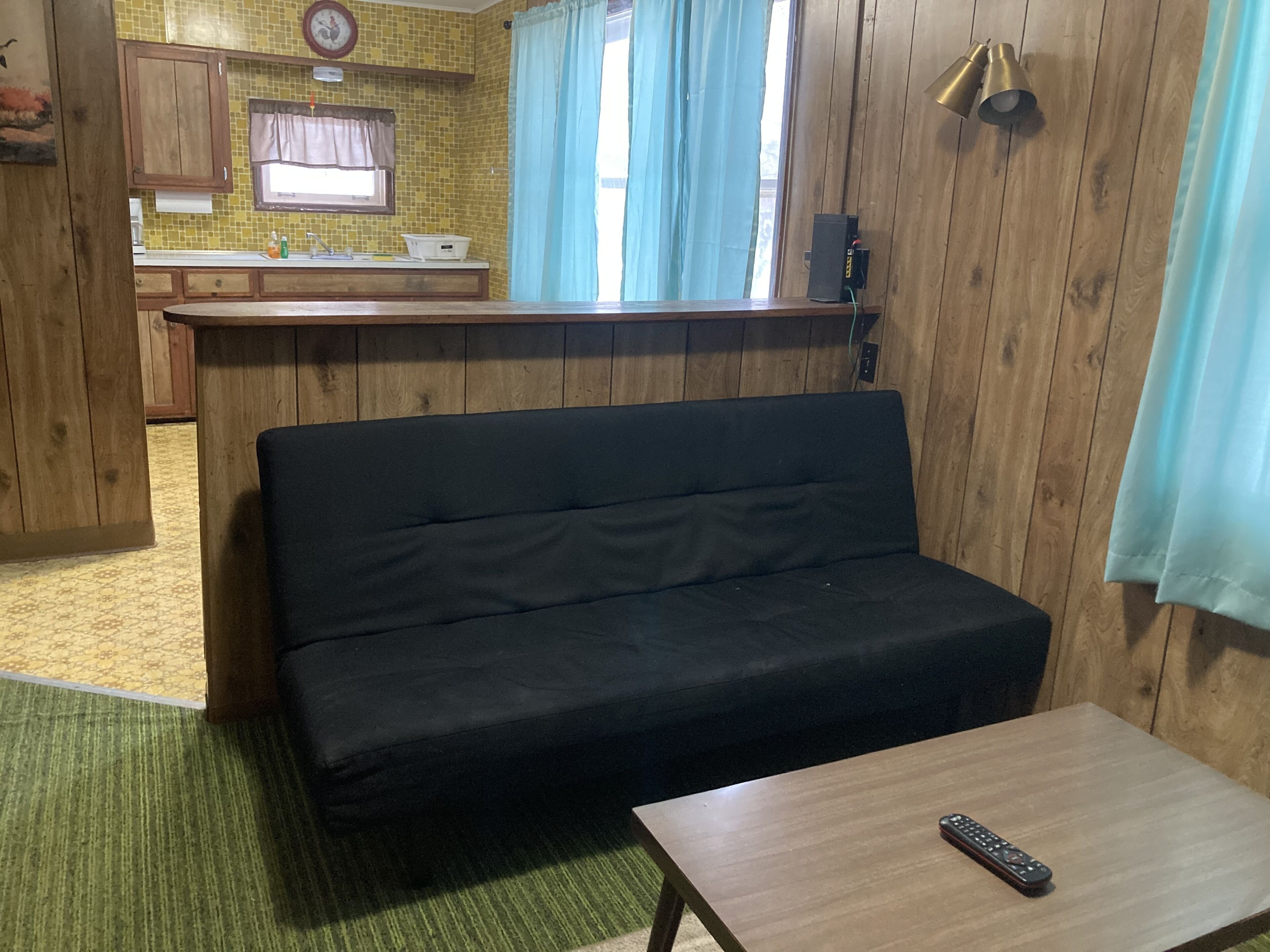 A room with a black sofa, a wooden table, a remote control, a kitchen area with wooden cabinets, a window with curtains, and a clock on the wall.