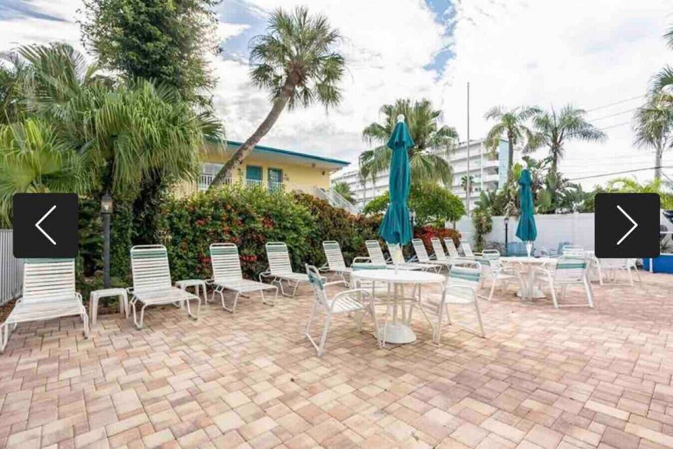 A patio area with white metal chairs and tables, surrounded by palm trees and a building with a blue roof.