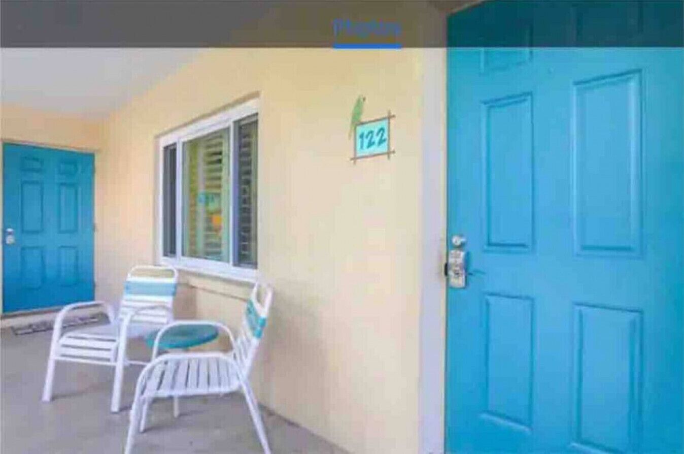 A blue door with a number sign, a window with blinds, and two white chairs outside.