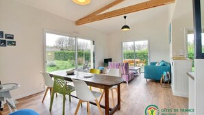 A spacious living area with a wooden dining table, chairs, and a sofa. There are large windows providing a view of the garden. - Le Grand Chemin - Ferel (FEREL)