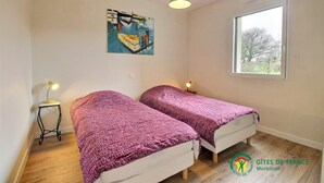 A bedroom with two beds, a painting on the wall, a window, and a small table with a lamp. - Le Grand Chemin - Ferel (FEREL)