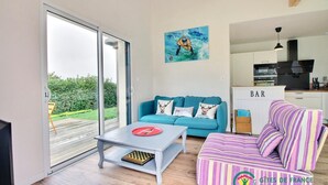 A modern living room with a teal sofa, a striped ottoman, and a coffee table with a red plate. A painting of a surfer is on the wall, and there's a kitchen area with a bar counter. - Le Grand Chemin - Ferel (FEREL)