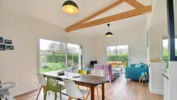 A spacious living area with a wooden dining table, chairs, and a sofa. There are large windows providing a view of the garden.
