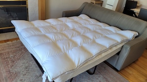 A sofa bed with a white cover, positioned in a room with a patterned rug, a wooden floor, and a small table with a lamp.
