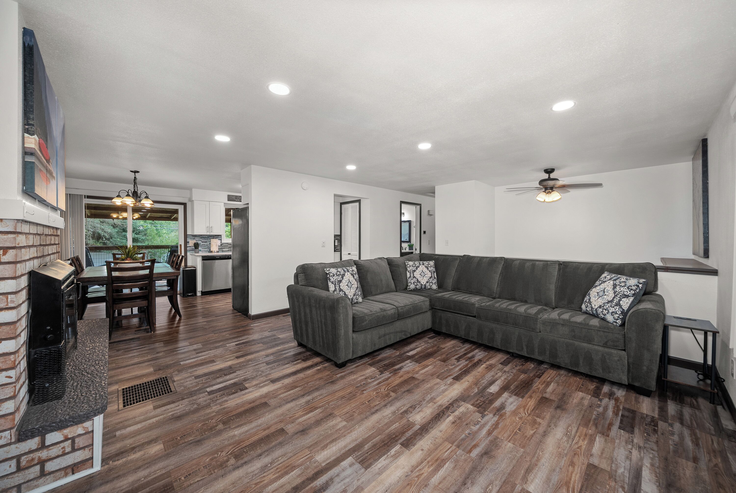 A spacious living room with a sectional sofa, a dining area with a brick fireplace, and a kitchen in the background.
