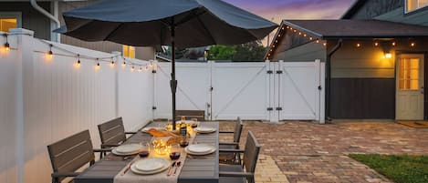 Un patio al aire libre con una mesa lista para comer, rodeada de sillas y una sombrilla.