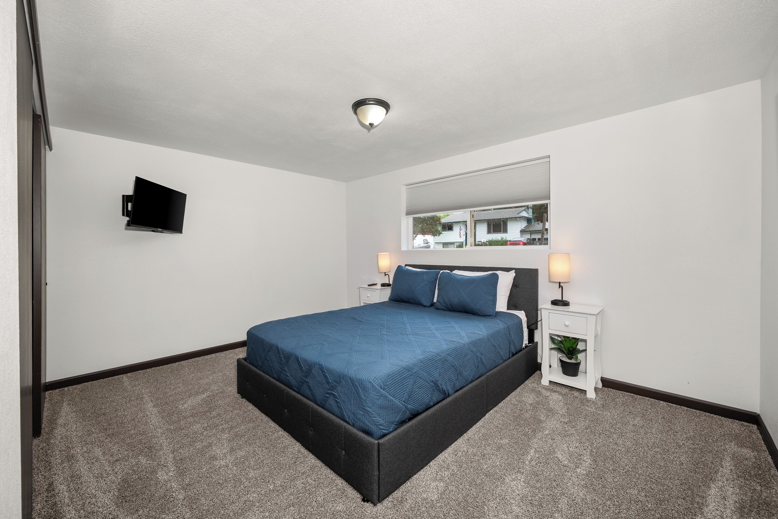 A hotel room with a bed, a television, a nightstand with a lamp, and a window with a view of the outside.