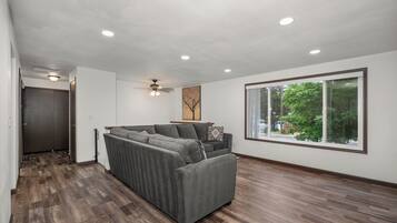 A modern living room with a sectional sofa, a ceiling fan, recessed lighting, and a large window with a view of greenery.