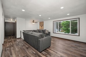 Un salon moderne doté d’un canapé d’angle, d’un ventilateur de plafond, d’un éclairage encastré et d’une grande fenêtre offrant une vue sur la verdure.
