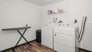 Una sala de lavandería con lavadora, secadora, tabla de planchar y estantes con productos de limpieza.