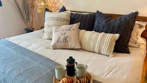 Uma cama bem arrumada com um travesseiro decorativo, uma bandeja com croissants e café, e uma atmosfera acolhedora com uma luminária e plantas.