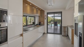 A modern kitchen with white cabinets, a built-in oven, and a window overlooking a pool. - Pool, sun and sea view - Villa das Quebradas (Funchal)