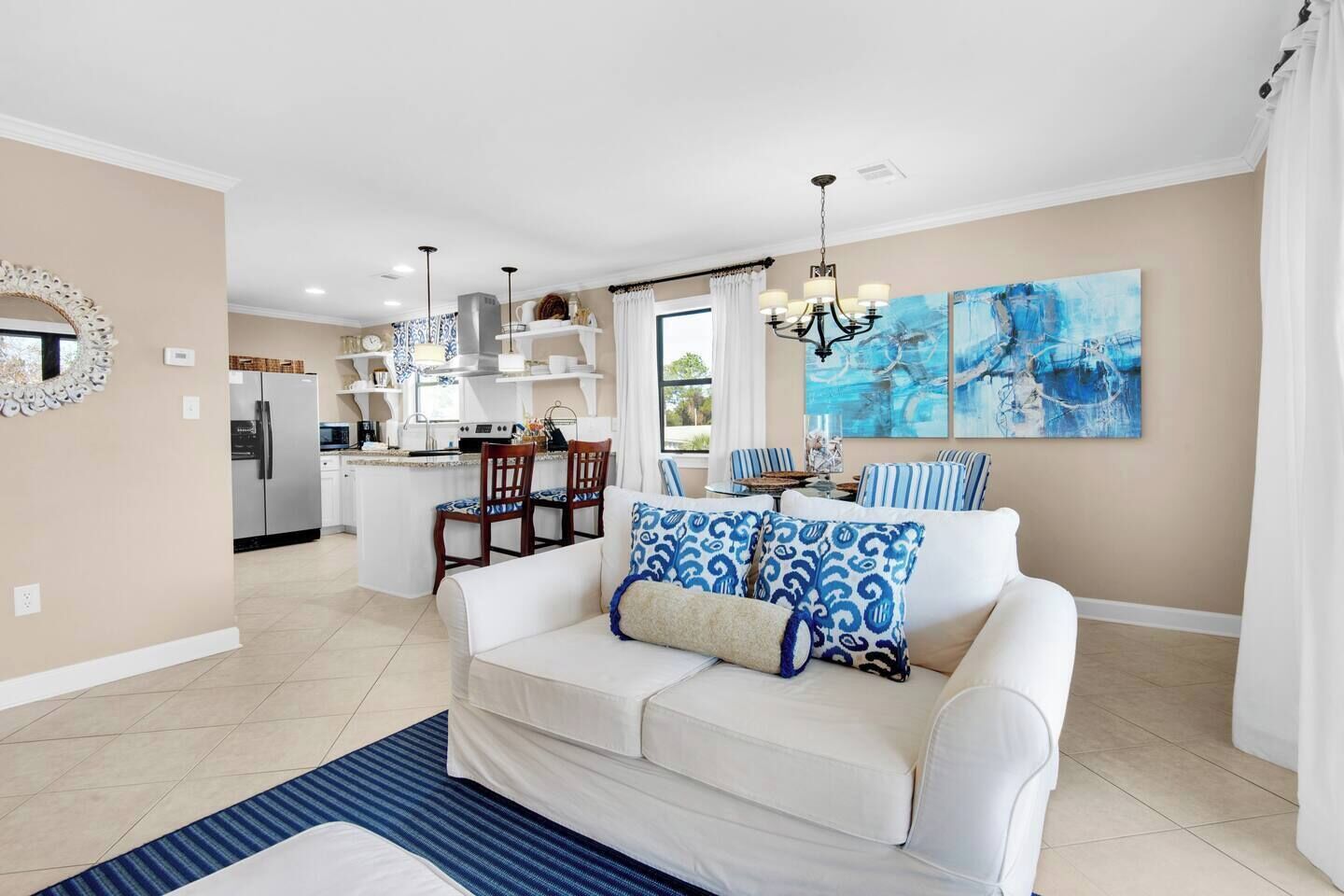 A modern living room with a white sofa, blue patterned cushions, a striped rug, and a dining area with striped chairs and a kitchen in the background.