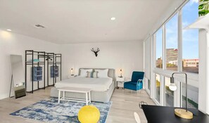 Ein modernes Schlafzimmer mit einem großen Bett, einem Schreibtisch, einem Stuhl und Blick auf die Stadt durch große Fenster.