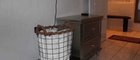 Una cesta de ropa pequeña con asa marrón sobre un taburete negro en una habitación con una puerta blanca, un banco de madera y un suelo de baldosas.