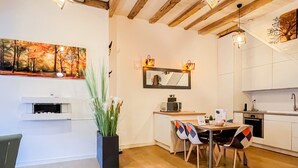 A modern kitchen with white cabinets, a dining area with a wooden table and chairs, a potted plant, and a painting on the wall. - Love island romantique rue carnot (Annecy)