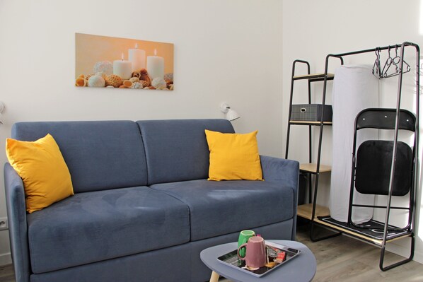 A blue sofa with yellow cushions, a small table with a magazine and two mugs, a bunk bed with a hanging rack, and a painting on the wall.