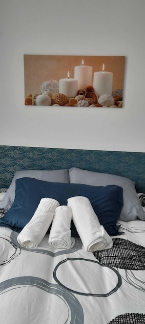 En snyggt bäddad säng med blå och vita kuddar, en vit pläd och hoprullade vita handdukar. Ovanför sängen hänger en inramad bild med motiv av ljus och snäckskal.