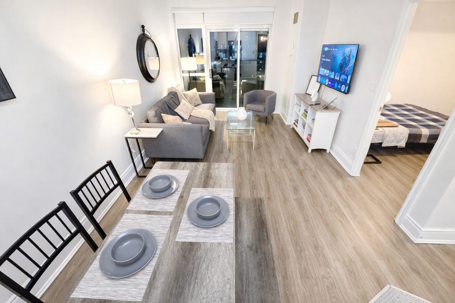 Una sala de estar moderna con un sofá, una mesa de comedor dispuesta con platos, un televisor y una cocina visible a través de una puerta de cristal.