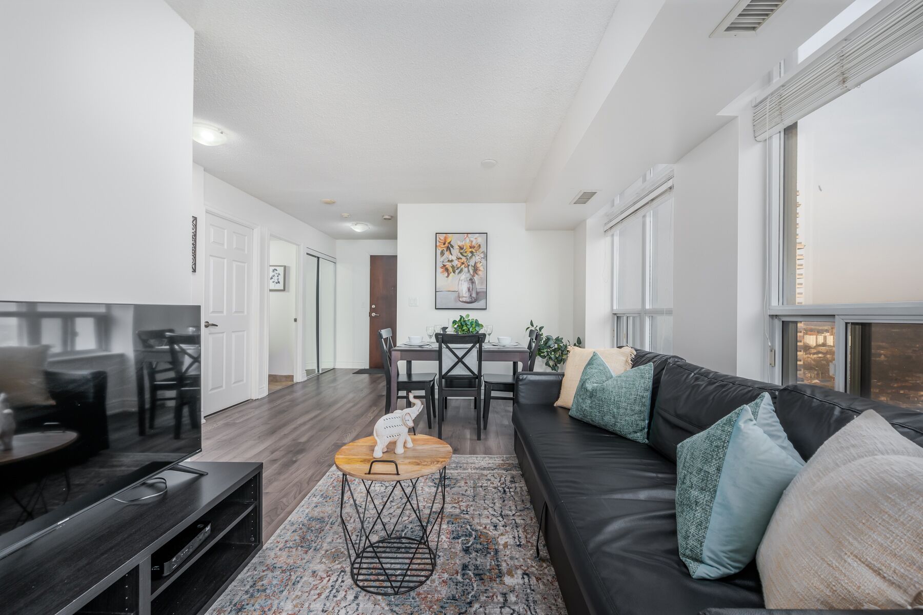 A modern living room with a black leather sofa, a coffee table, and a TV stand. There is a dining area with a table and chairs, and a painting on the wall.
