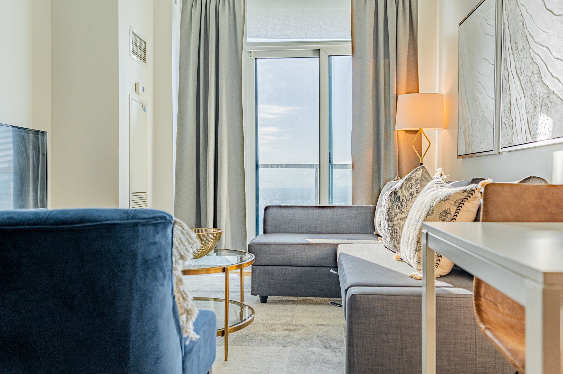 Una habitación de hotel moderna con un sofá gris, un sillón azul, una mesita redonda y una lámpara.